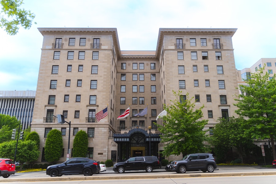 The exterior of the historic Jefferson Hotel on a sunny day with flags, trees and parked cars out front. 