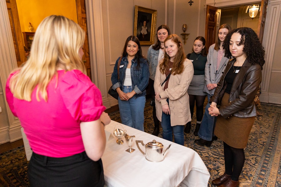 A group of women talking to a woman in a pink shirt about a tea set in an historic house
