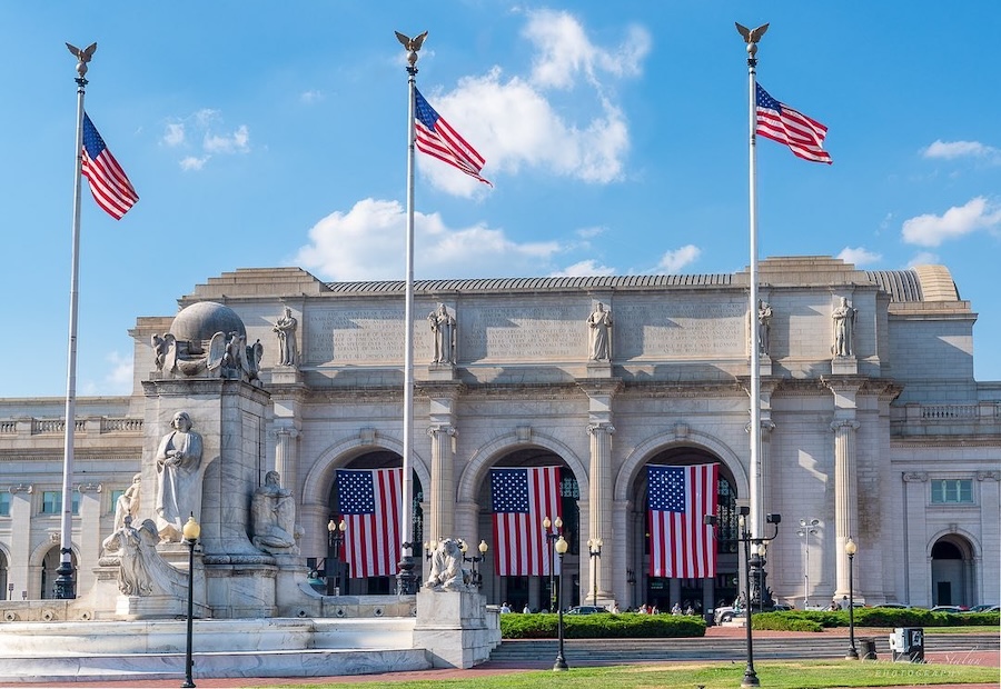 The exterior of Union Station in Washington, DC is adorned with large American flags and patriotic statues under a bright blue sky.