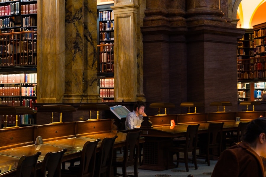 A man studies at a lamp-lit desk in the Library of Congress reading room surrounded by towering bookshelves and marble columns.