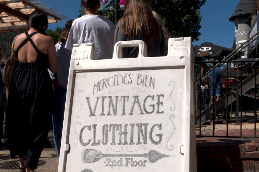 A sign for Mercedes Bien vintage clothing stands in front of a group of people waiting outside.