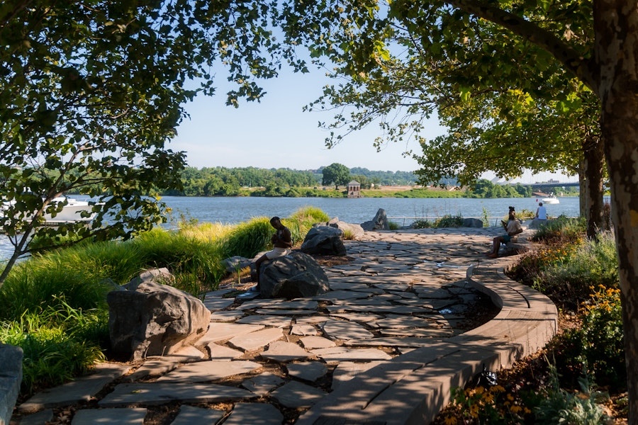 A stone pathway curves along the Anacostia River with people relaxing under shady trees at Yards Park.