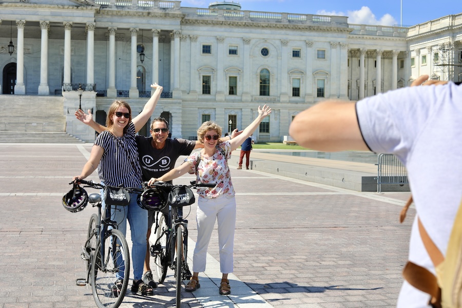 Three cyclists pose for a photo in front of the U.S. Capitol during a bike tour, raising their arms and smiling.