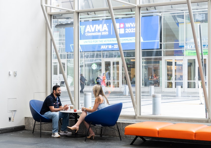 Two people sit and talk over drinks in a bright lobby area with large windows showing signage for the AVMA 2025 convention outside.