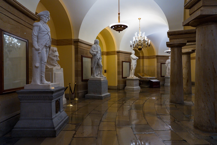Marble statues line the arched corridors of the U.S. Capitol Crypt beneath the Rotunda, illuminated by chandeliers.
