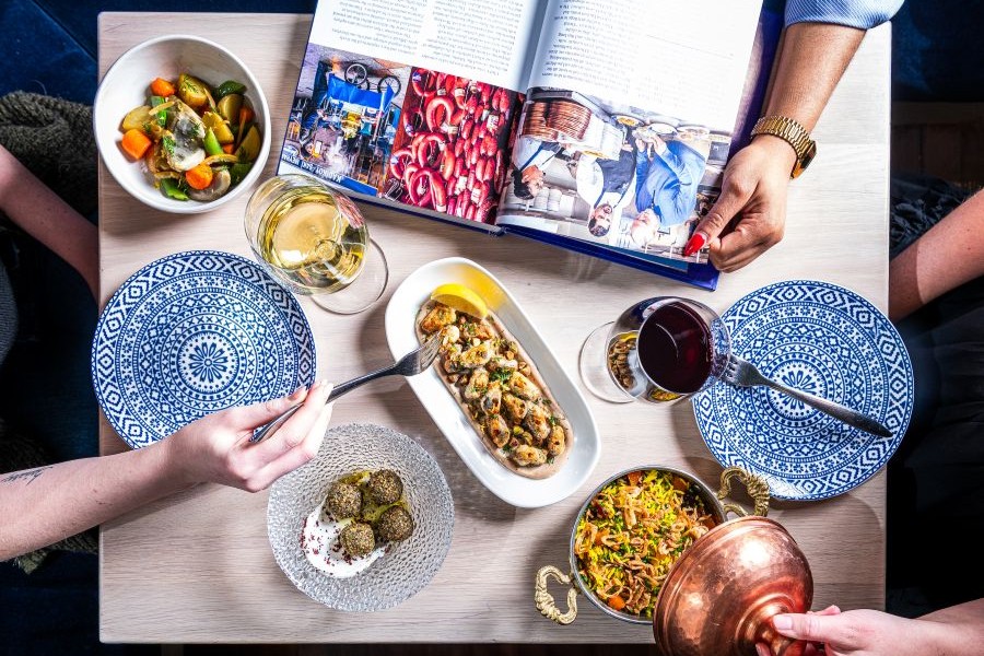 A brightly lit tabletop displays several colorful Mediterranean-style dishes, patterned blue plates, and hands reaching in to serve and share food beside an open cookbook.
