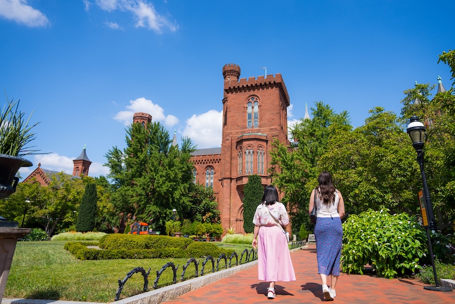 Two women walk along a garden path toward the Smithsonian Castle surrounded by lush greenery.