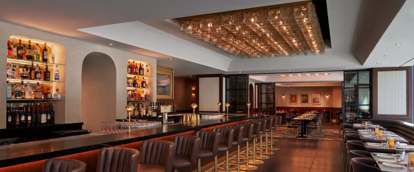 A stylish restaurant and bar with a long, sleek counter lined with leather barstools, illuminated liquor shelves, and a warmly lit dining area with modern decor in the background.