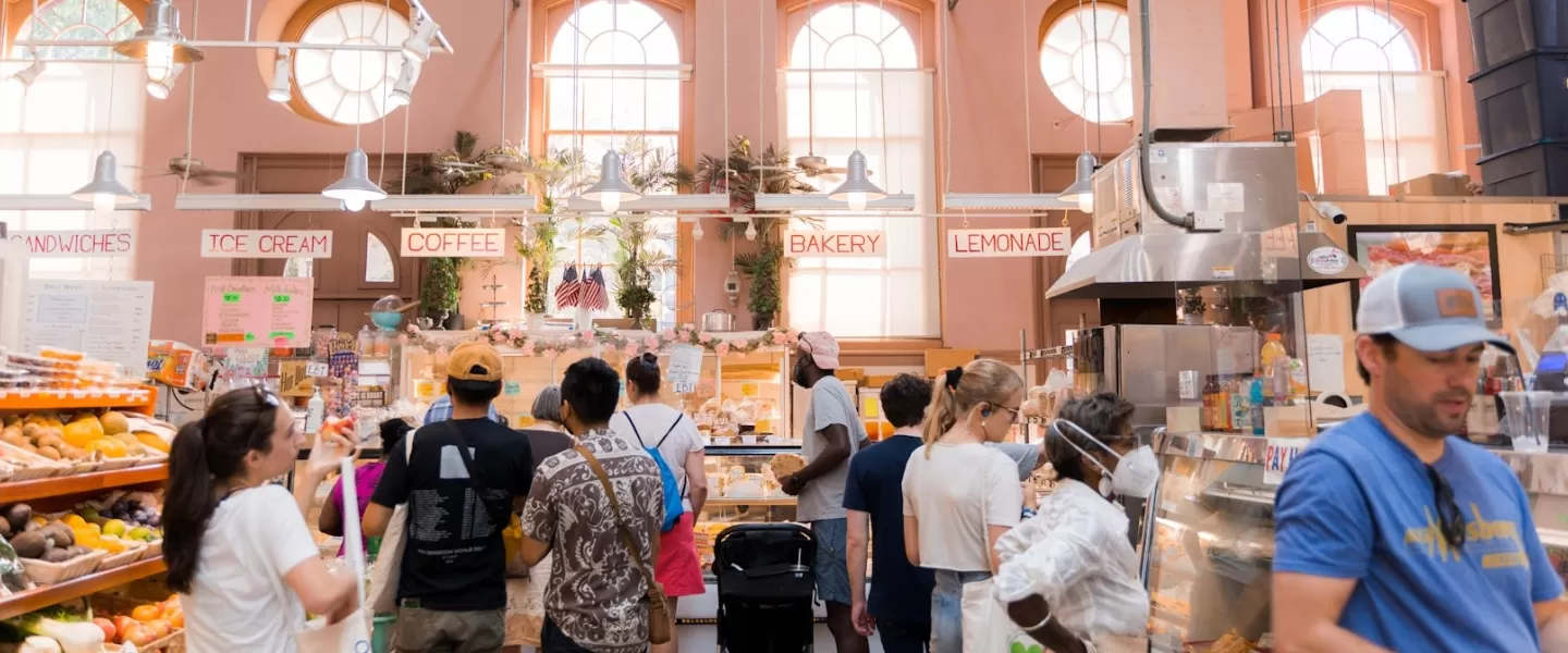 Shoppers queue at a brightly lit indoor bakery and deli inside Eastern Market, with signs for sandwiches, coffee and lemonade.