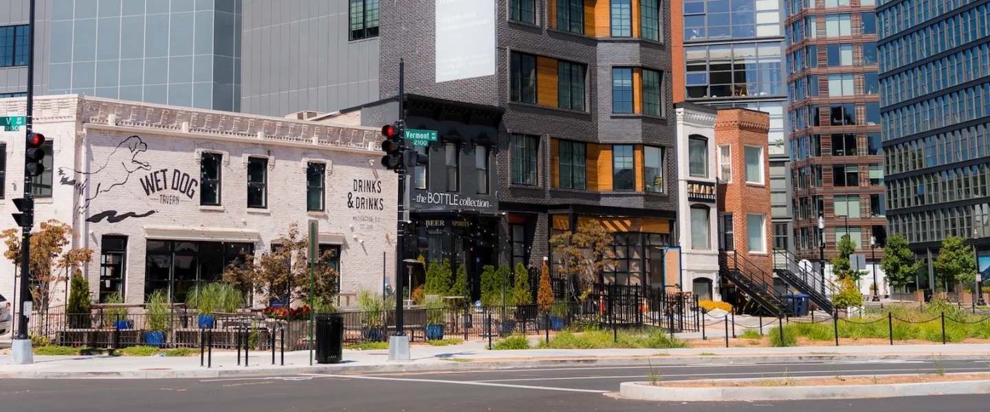 Wet Dog Tavern and nearby storefronts line a sunny street in Shaw, framed by modern buildings.