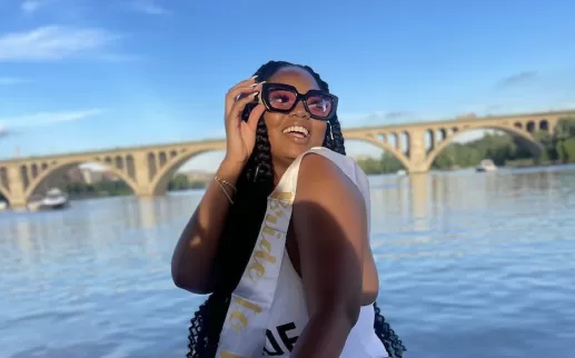 A smiling woman wearing large sunglasses and a 'Bride to Be' sash poses near a river with an arched bridge in the background.
