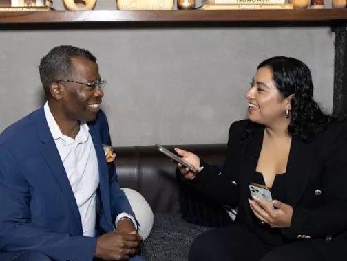 This image shows two people sitting on a couch, engaged in a friendly and professional conversation. The woman on the right is holding a phone, possibly conducting an interview. Both are smiling, suggesting a positive and engaging discussion. The setting appears casual yet professional, possibly a media or networking event.