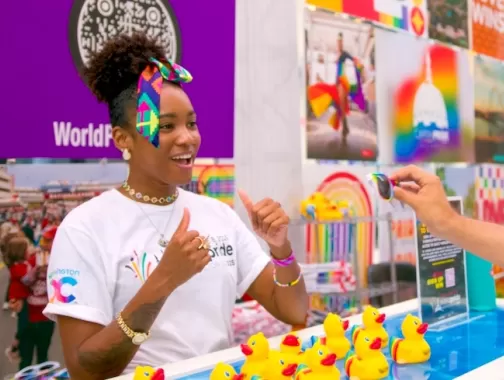 A young woman with a rainbow bow in her hair plays a game with an attendee at an event booth.