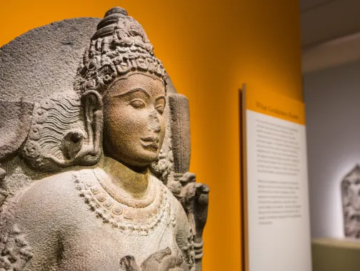 A carved stone Hindu figure is displayed in a museum gallery with warm yellow lighting in Washington, DC.