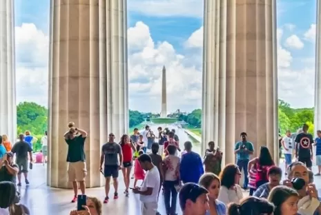 A crowd of visitors inside the Lincoln Memorial with a view of the Washington Monument beyond the columns.