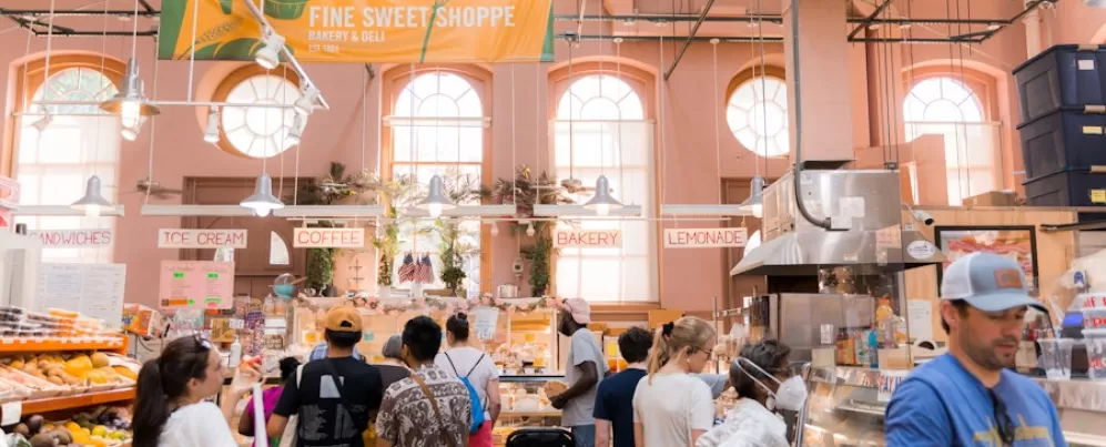 Shoppers line up at a bakery stall inside Eastern Market. The bright and airy space is filled with natural light, with signs for coffee, ice cream, and bakery items hanging above.